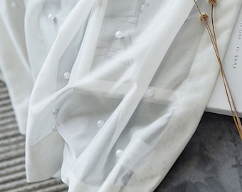 Puede incluir: Cortina blanca transparente con pequeñas perlas blancas. La cortina está drapeada sobre un libro con texto en un idioma extranjero.