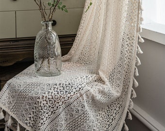 Puede incluir: Un mantel de crochet blanco con un delicado patrón de encaje y flecos de borla. El mantel está drapeado sobre una silla y tiene una estética vintage y rústica.