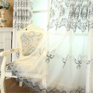 Könnte beinhalten: Ein cremefarbener Stuhl mit gemustertem Sitz und Rücken steht vor einem Fenster mit transparenten Vorhängen. Die Vorhänge und der Stuhl weisen verzierte graue und blaue Blumenmuster auf, die eine elegante, klassische Ästhetik schaffen.