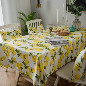 May include: White dining table with a lemon print tablecloth. The table is set with white chairs and cushions with the same lemon print.