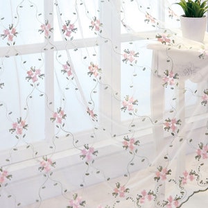 May include: White sheer curtains with a delicate floral pattern in pink and green. The curtains are hung in a window with a white frame.