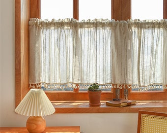 May include: A white sheer valance with a fringe trim hangs in a window with wood trim. A small potted plant sits on the windowsill next to a closed book.