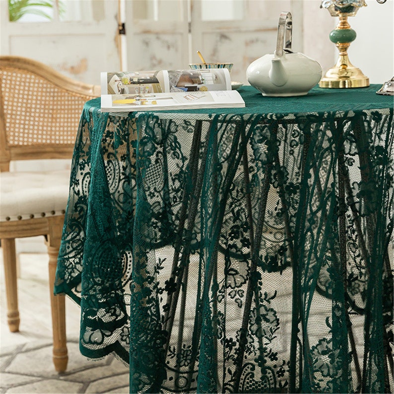 May include: A green lace tablecloth with a floral pattern. The tablecloth is draped over a round table with a book and a teapot on top.