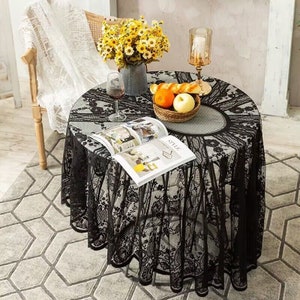May include: Black lace tablecloth with a delicate floral pattern. The tablecloth is round and has a scalloped edge. It is laid on a table with a vase of yellow flowers, a book, and a bowl of fruit.