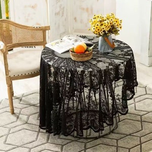 May include: Black lace tablecloth with a scalloped edge, draped over a round table with a book and a bowl of fruit on top. The table is set in a room with a white wall and a wicker chair.