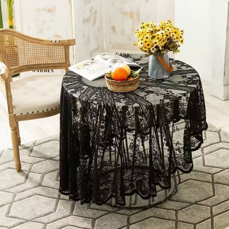 May include: Black lace tablecloth with a scalloped edge, draped over a round table with a book and a bowl of fruit on top. The table is set in a room with a white wall and a wicker chair.