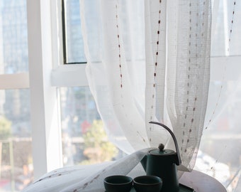 May include: A white sheer curtain with brown embroidered dots hangs in front of a window. A green teapot and two green teacups sit on a wooden table in front of the curtain.