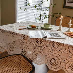 May include: A cream lace tablecloth with a circular design covers a wooden table. Green plates, a bowl, and a magazine are on the table. Two lit candlesticks and a wooden serving board are also present.