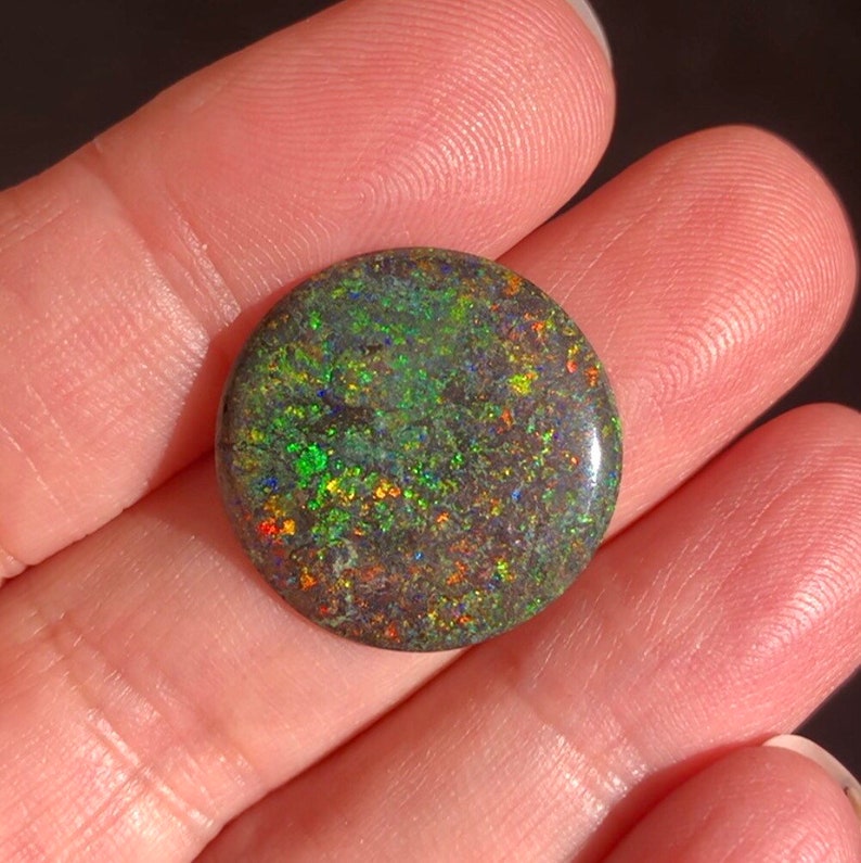 May include: A round, dark green opal cabochon with iridescent flashes of blue, red, and yellow.