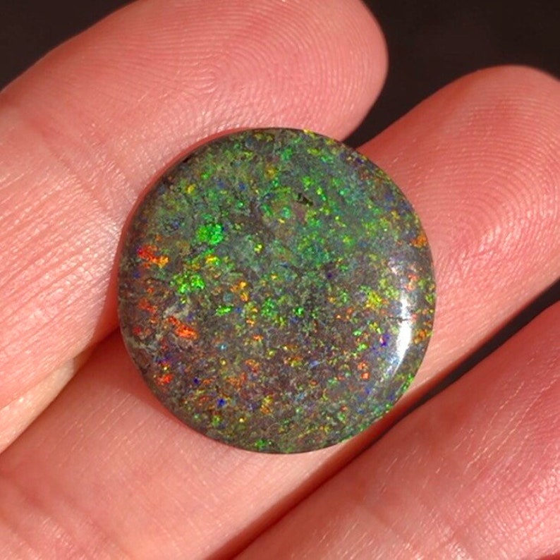 May include: A round, dark green opal cabochon with iridescent flashes of blue, red, and green.