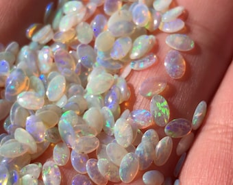 May include: A collection of oval-shaped white opal gemstones with iridescent rainbow colors. The opals are scattered on a hand.
