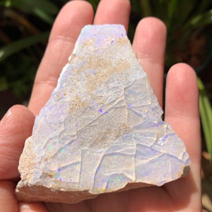 May include: A rough, white opal with iridescent blue and green flashes. The opal is a natural stone and has a rough, irregular shape.