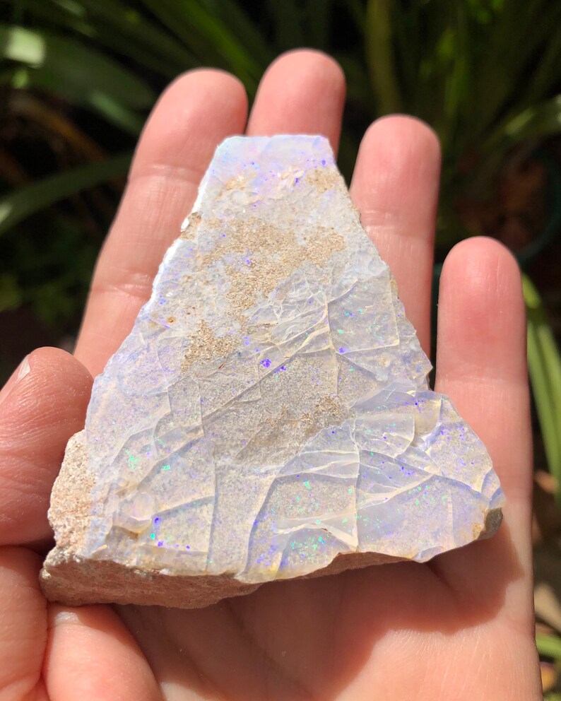 May include: A rough, white opal with iridescent blue and green flashes. The opal is a natural stone and has a rough, irregular shape.