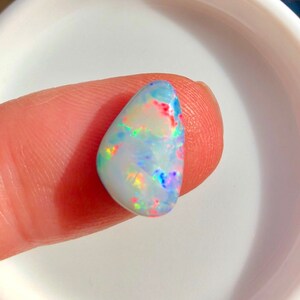 Puede incluir: Una piedra preciosa de ópalo pulida, de forma ovalada. El ópalo muestra un color base blanco lechoso con destellos de rojo, azul, verde y amarillo. La gema se sostiene entre dos dedos, mostrando su juego de colores iridiscente.