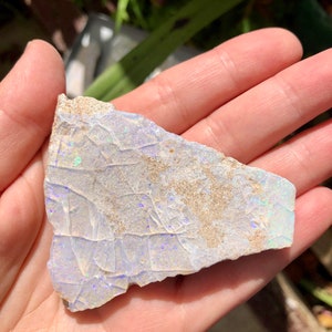 May include: A piece of rough opal with a white base and iridescent blue and green flashes. The opal is irregular in shape and has a rough, textured surface.