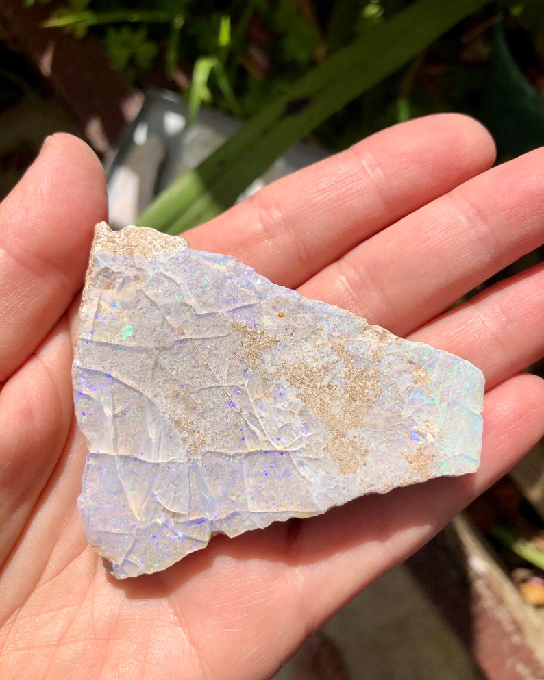May include: A piece of rough opal with a white base and iridescent blue and green flashes. The opal is irregular in shape and has a rough, textured surface.