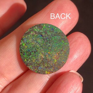 May include: A round, green opal cabochon with a dark, iridescent surface. The back of the opal is visible, showing a dark, green, and brown pattern.