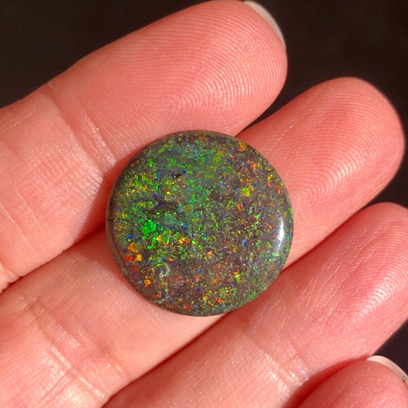 May include: A round, dark green opal cabochon with flashes of red, orange, and blue.