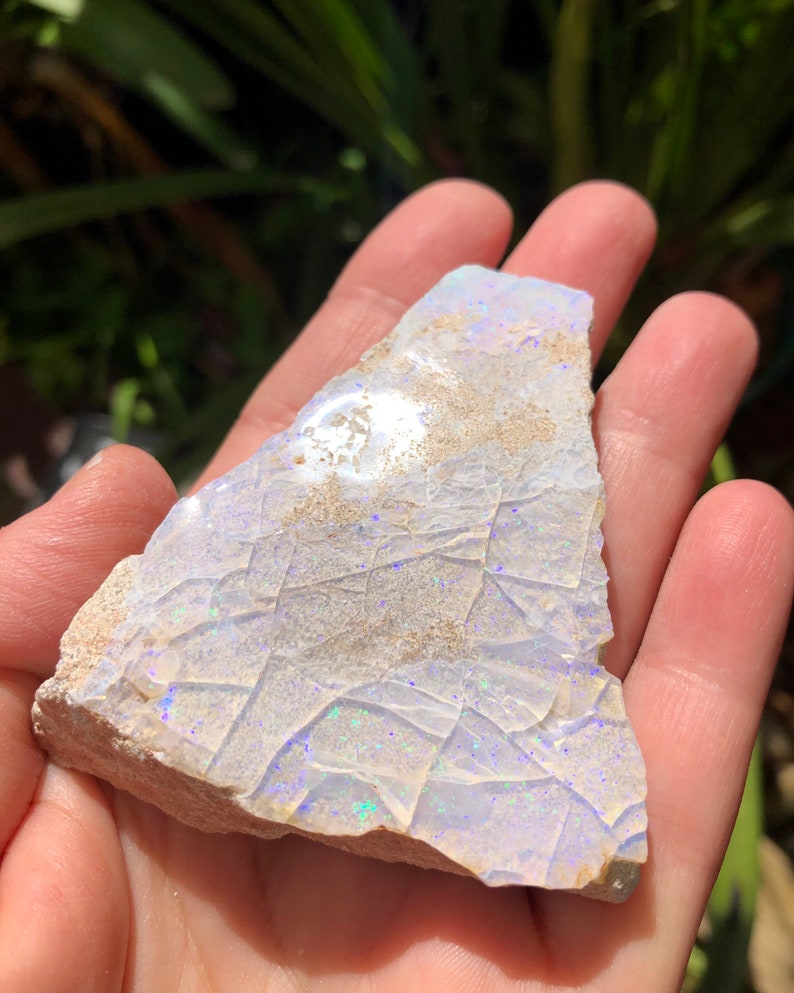 May include: A rough, white opal with iridescent blue and green flashes. The opal is a natural stone with a rough, irregular shape.