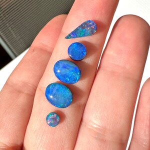 May include: A selection of polished opals in different shapes and sizes. The opals exhibit a vibrant play-of-colour, with flashes of blue, green, and purple. The gemstones are oval, round, and teardrop-shaped, showcasing their unique internal patterns.