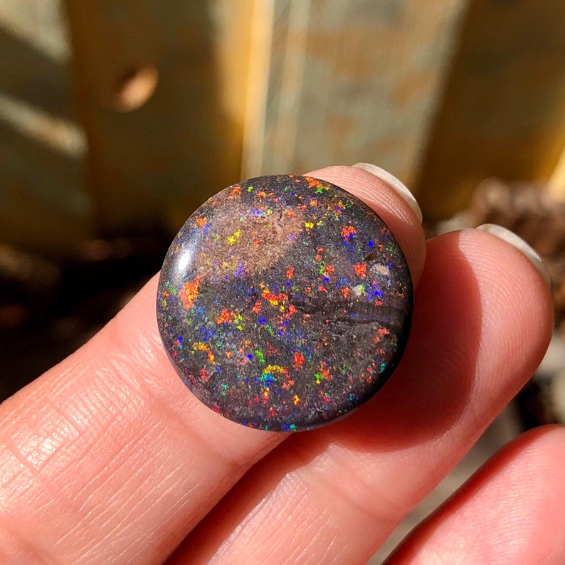 May include: A round, black opal cabochon with a dark, iridescent surface. The opal has a speckled pattern of red, orange, green, and blue.