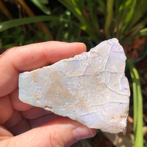 May include: A white opal with iridescent blue and purple flashes. The opal is rough and has a natural, irregular shape.