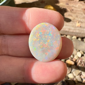 May include: A white opal cabochon with iridescent rainbow flashes. The opal is oval shaped and has a smooth, polished surface.