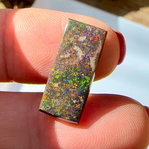 Puede incluir: Una piedra preciosa de ópalo rectangular de color marrón oscuro con una mezcla de colores verde, azul y rojo.