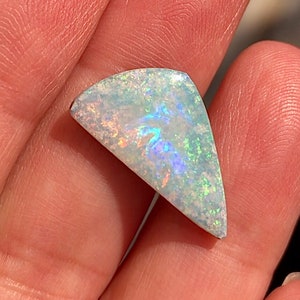 May include: A white opal gemstone with iridescent blue and green flashes. The opal is cut in a triangular shape and has a rough, textured surface.