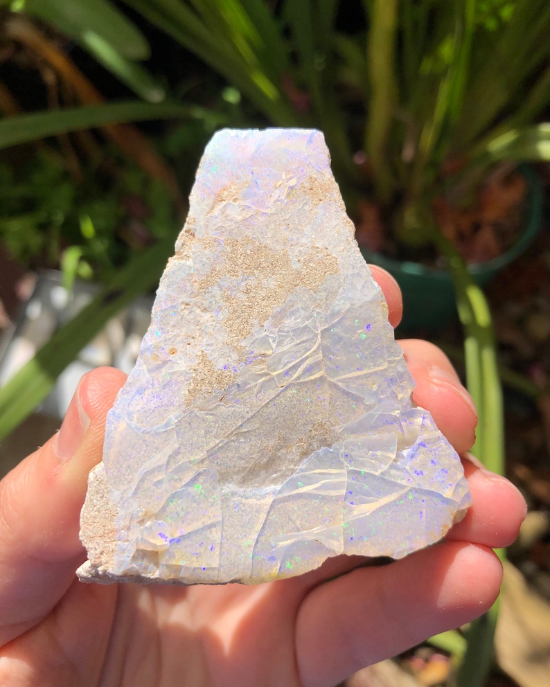 May include: A rough, white opal with iridescent blue and green flashes. The opal is a natural stone with a rough, irregular shape.
