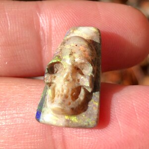 May include: A carved opal skull pendant with iridescent green, blue, and yellow colours. The skull is detailed with eye sockets, a nose, and a mouth.