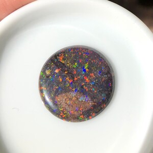 May include: A round black opal cabochon with a dark background and colorful flashes of red, orange, yellow, green, blue, and purple.
