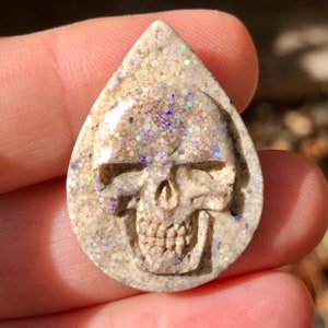 May include: A white opal cabochon carved into a skull shape. The opal has a sparkly, iridescent finish with a light brown base.