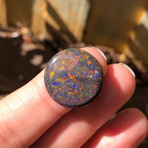May include: A round, black opal gemstone with a dark, mottled surface and flashes of iridescent color, including blue, green, orange, and red.