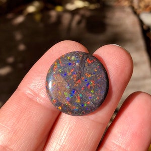 May include: A round, dark gray opal cabochon with a black matrix and iridescent flashes of red, orange, yellow, green, blue, and purple.