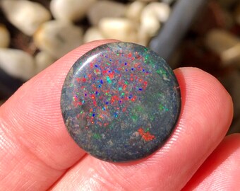 May include: A round, dark gray opal stone with vibrant red, blue, and green flecks. The polished surface of the gemstone reflects light, creating a sparkling effect. The opal is held in a hand, showcasing its natural beauty.