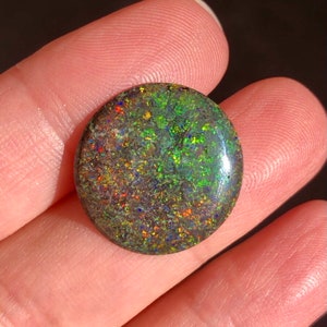 May include: A round, green, black, and red opal cabochon gemstone with a high polish. The opal has a dark body tone with flashes of colour.