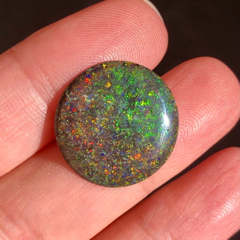 May include: A round, green, black, and red opal cabochon gemstone with a high polish. The opal has a dark body tone with flashes of colour.