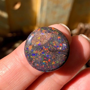 May include: A round, black opal cabochon with a dark, iridescent surface. The opal has a speckled pattern of red, orange, green, blue, and purple.