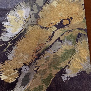May include: Close-up of a textile with a dark purple background and a pattern of gold, silver, and green brushstroke-like designs. The fabric has a textured appearance, with the gold elements appearing to shimmer.