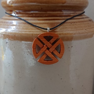 Puede incluir: Un collar con un colgante de madera de color marrón, con un diseño de nudo intrincado. El colgante cuelga de un cordón negro.