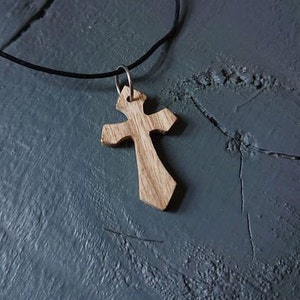 Puede incluir: Un colgante de cruz de madera en un collar de cordón negro. La cruz está tallada con un diseño detallado.