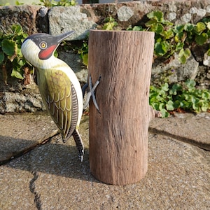 Puede incluir: Escultura de madera de un pájaro carpintero verde con cabeza roja y pico negro posado en un tronco de madera marrón. El pájaro tiene marcas detalladas de plumas. El tronco tiene una superficie rugosa.