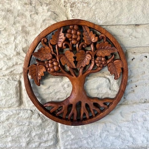 Peut inclure: Décoration murale ronde en bois représentant un arbre sculpté. L'arbre a un tronc, des racines, des feuilles et des grappes de raisin détaillés. La sculpture en bois brun foncé est posée sur un mur de briques blanches.