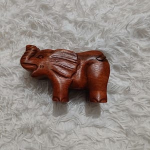 Puede incluir: Una figurita de elefante de madera con un acabado marrón. El elefante está tallado en posición de pie con la trompa levantada.