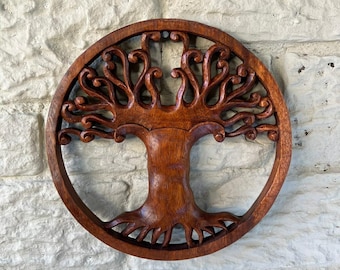 May include: A round, wooden wall hanging featuring a carved tree of life design. The tree has a thick trunk, detailed roots, and swirling branches within a circular frame. The wood has a rich, warm brown color, and the background is a textured, light-colored stone wall.