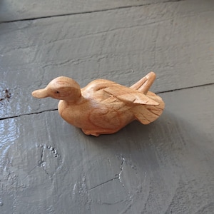 May include: A wooden carving of a duck with a light brown color. The duck is lying on its side with its wings spread out.