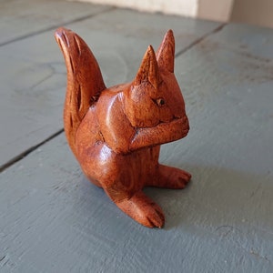 May include: A detailed wooden squirrel figurine, carved from a rich brown wood. The squirrel is depicted in a seated position with its tail arched upwards. The carving showcases the texture of the wood, highlighting the animal's features.