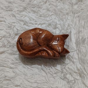 May include: A wooden puzzle box carved in the shape of a sleeping cat. The box is made of dark brown wood and has a detailed carving of the cat's features.
