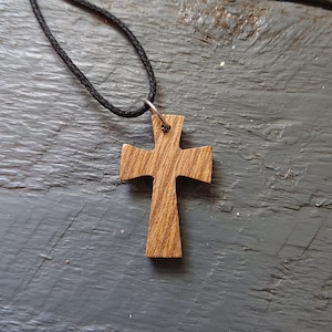Può includere: Un ciondolo a forma di croce in legno su una collana con cordoncino nero. La croce è in legno marrone chiaro con una venatura.